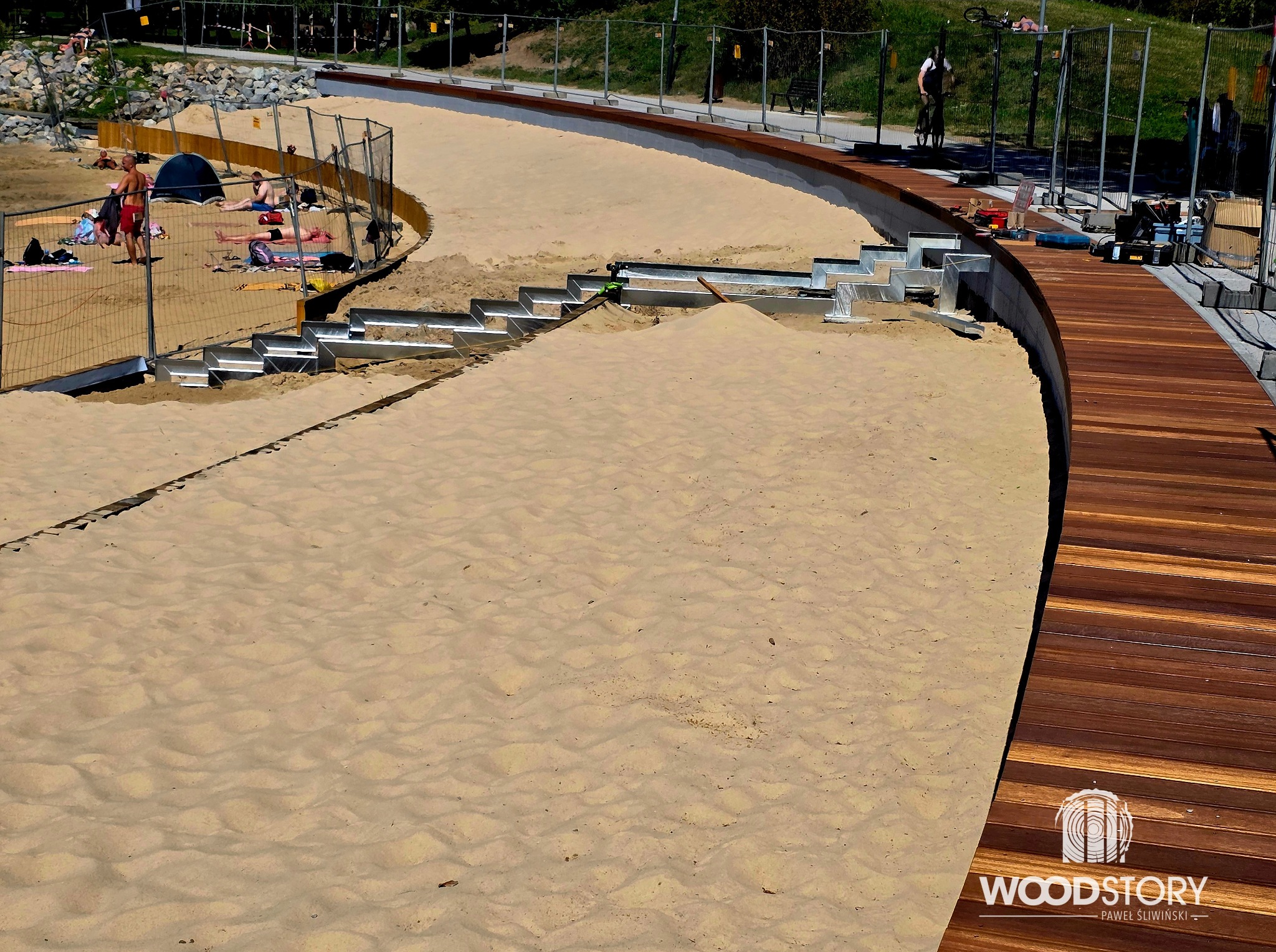 Budowa drewnianego tarasu na plaży z metalowymi schodami i widocznym piaskiem. W tle osoby odpoczywające na plaży, całość ogrodzona siatką.