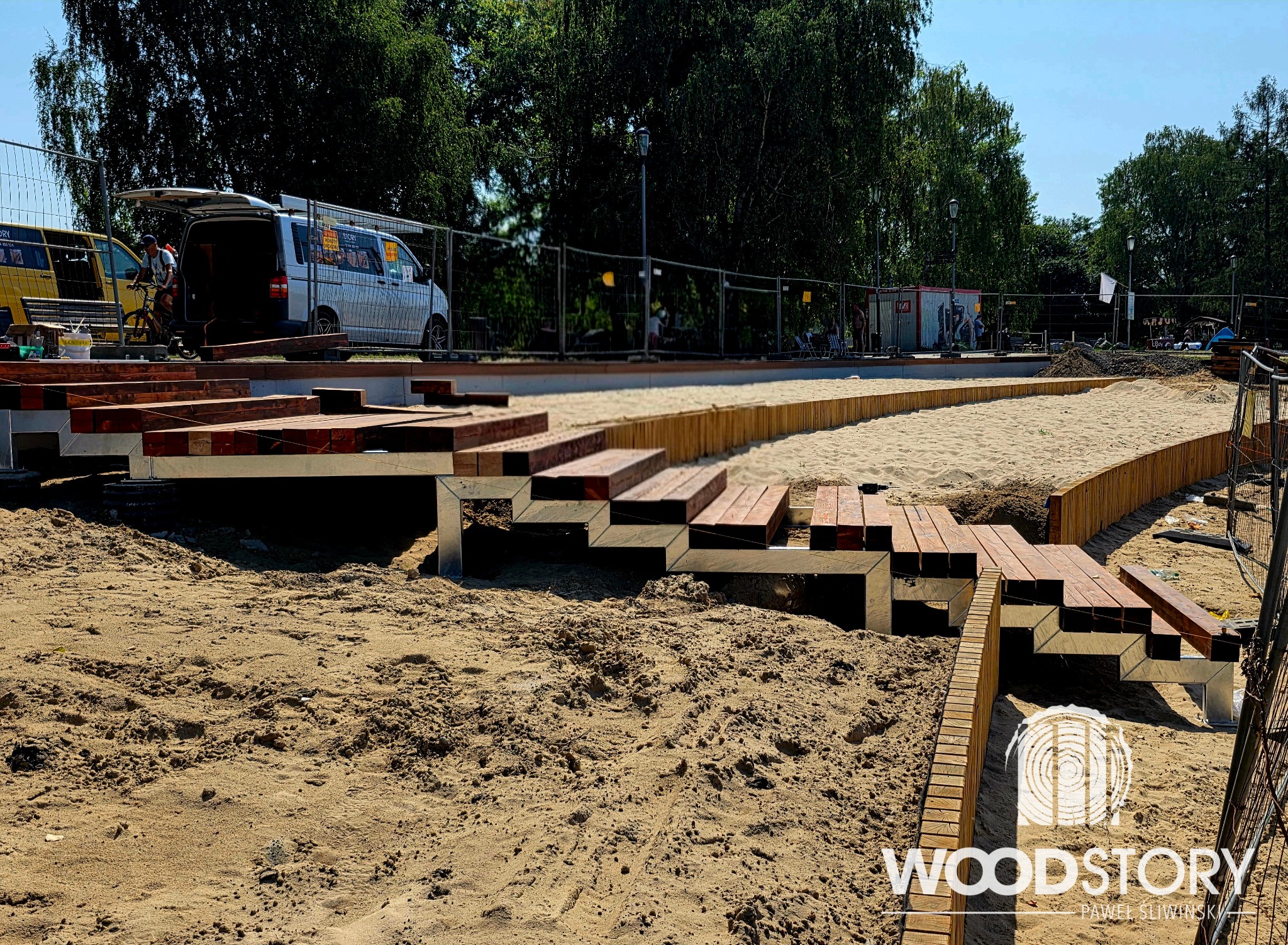 Budowa drewnianego tarasu schodkowego na piaszczystym podłożu. Widoczne elementy konstrukcyjne i etap prac. W tle pojazdy i zieleń.