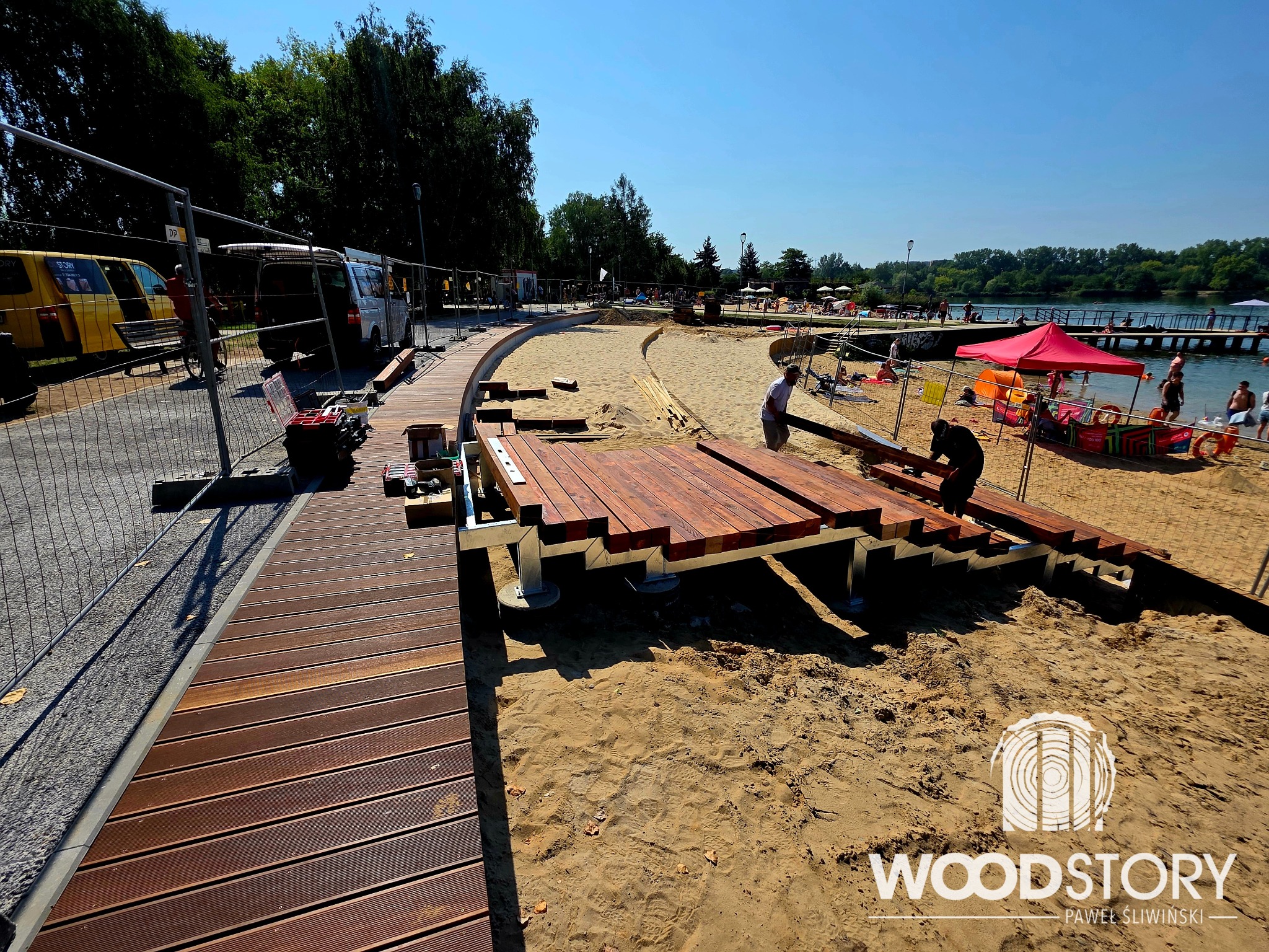Budowa drewnianego tarasu na plaży z widokiem na jezioro. W tle osoby odpoczywające na piasku, a w pierwszym planie pracownicy montujący konstrukcję.