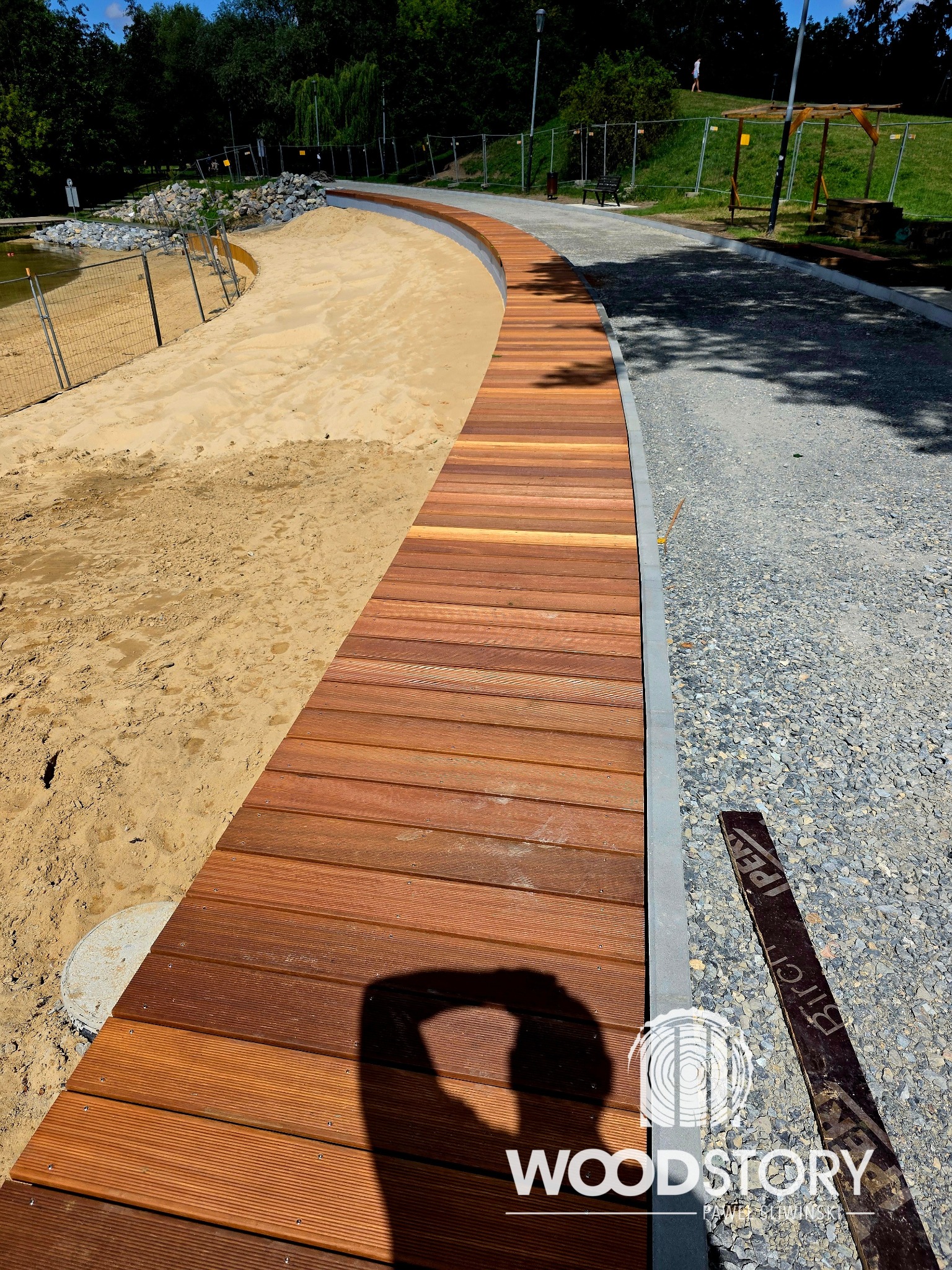 Zakrzywiony chodnik z desek na plaży, z widocznym logo firmy Woodstory. Deski w ciepłym odcieniu brązu, obok piasek i żwir. Metalowe obrzeże. Cień osoby robiącej zdjęcie.