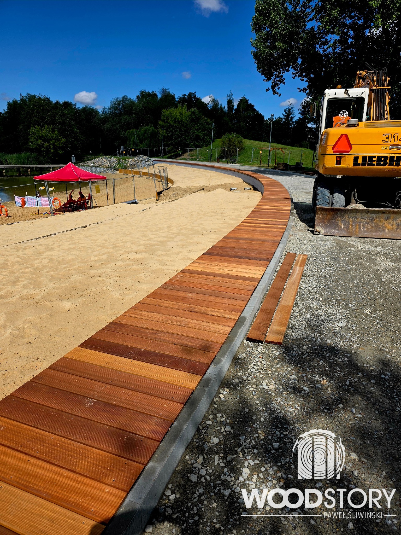Zakrzywiony drewniany chodnik z desek na plaży, obok piasku i żwiru, z koparką Liebherr w tle. Widoczny znak wodny Woodstory.