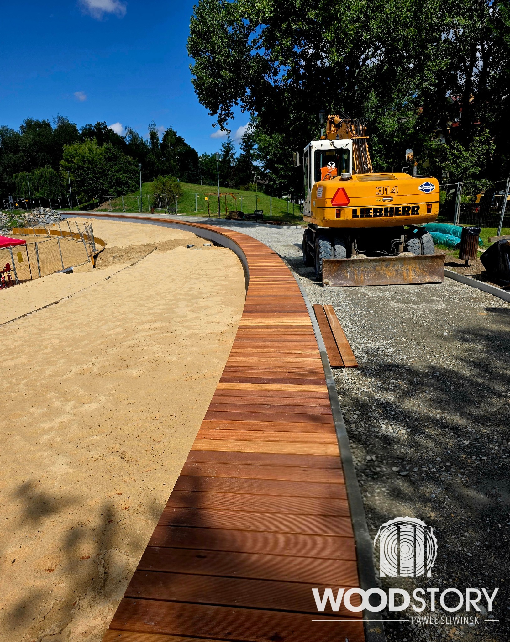 Zakrzywiony chodnik z desek w parku, obok piasku i utwardzonej ścieżki. W tle maszyna budowlana i zieleń. Widoczne logo firmy Woodstory.