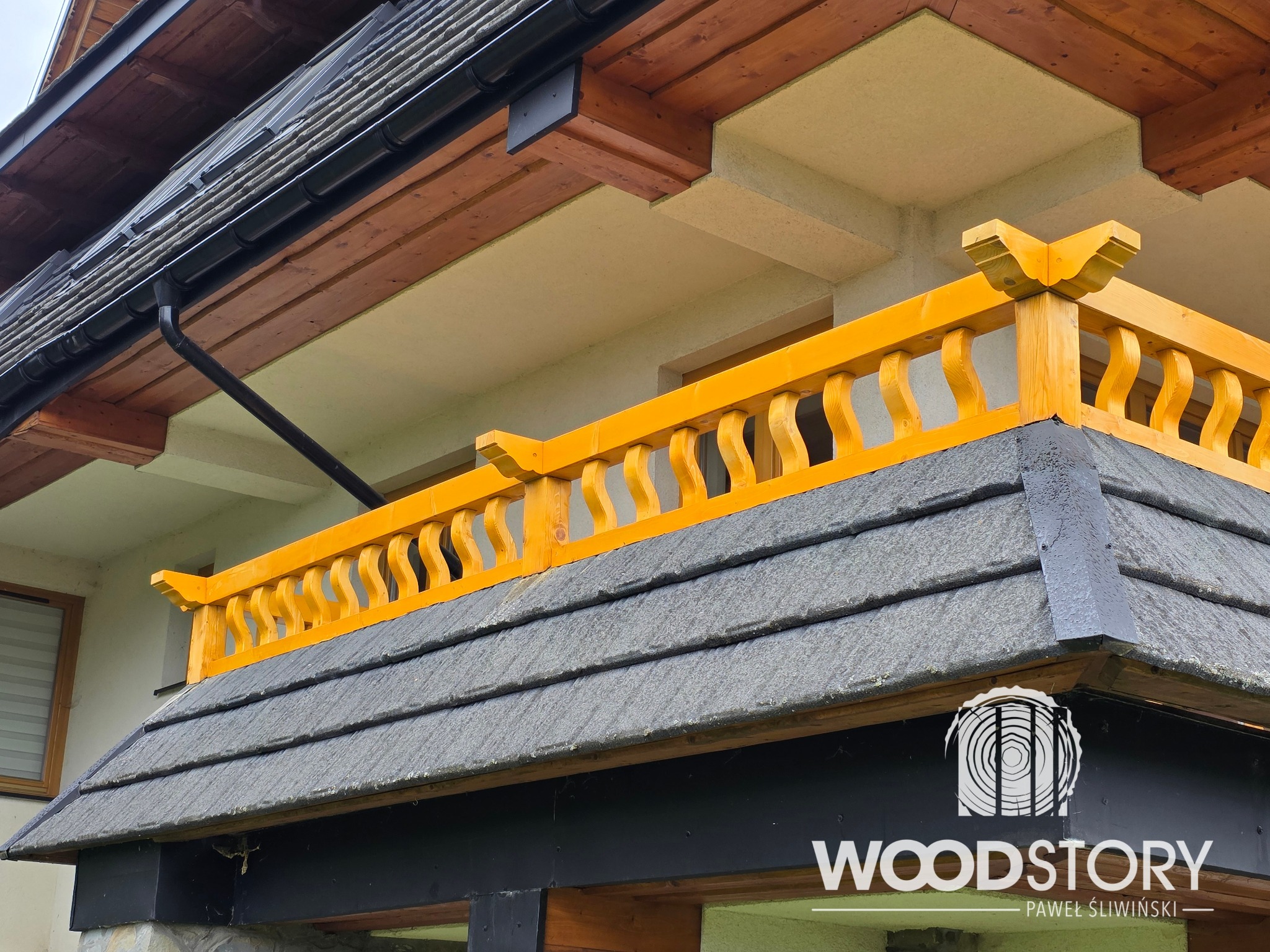 Żółta, drewniana balustrada z dekoracyjnymi wycięciami, zamontowana na zadaszeniu budynku z szarymi dachówkami. Widoczny fragment elewacji i logo firmy.