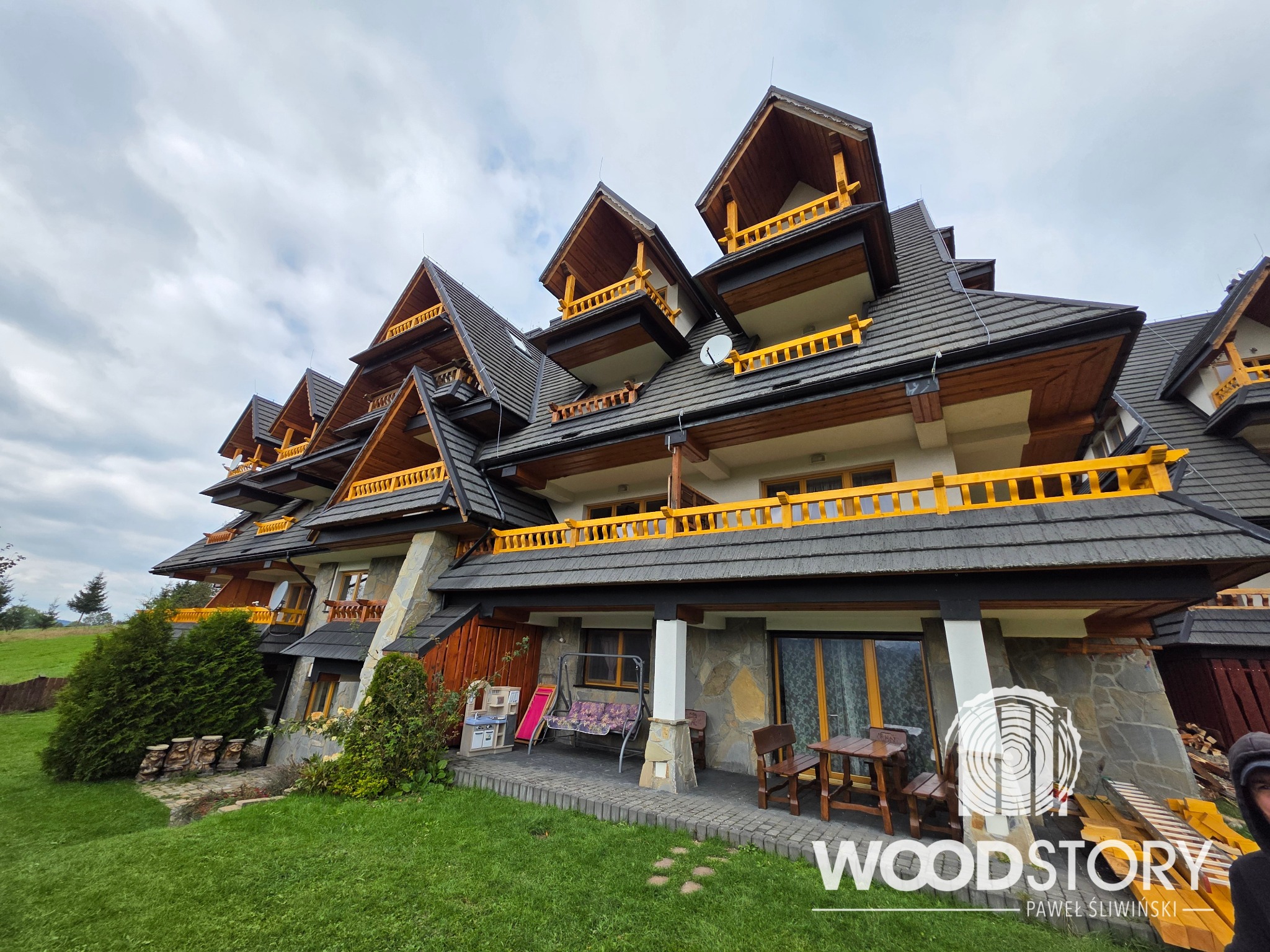 Rozległy dom w stylu góralskim z drewnianymi balkonami i balustradami w kolorze żółtym, widok z poziomu trawnika, pochmurne niebo w tle. Charakterystyczna architektura.