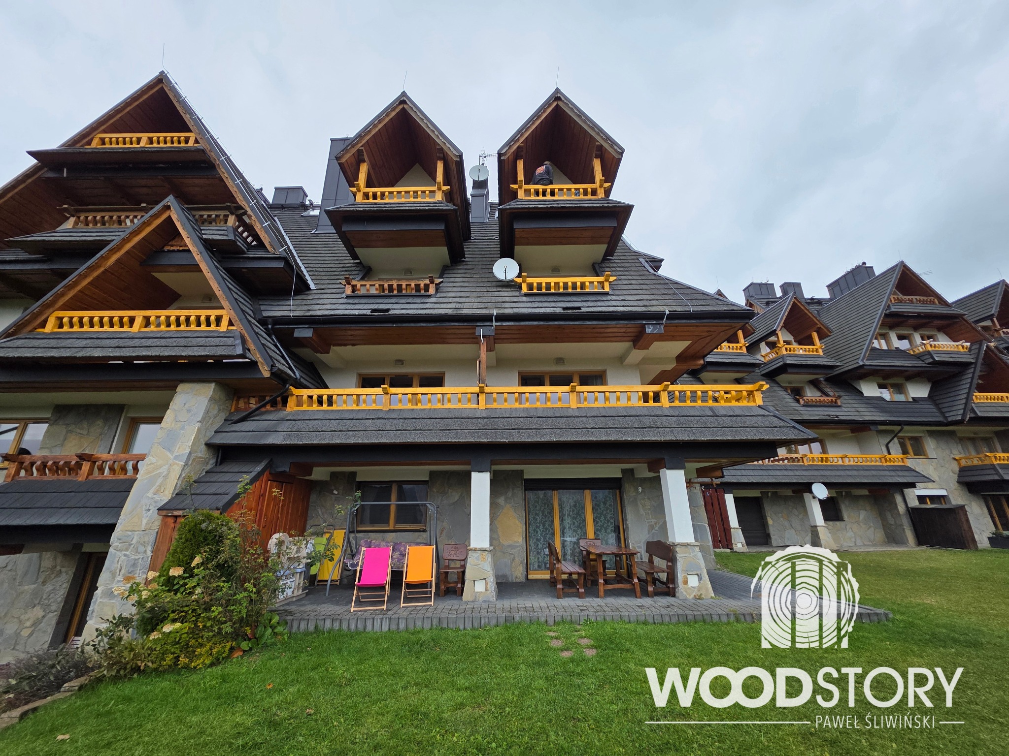 Kompleks budynków w stylu góralskim z drewnianymi balkonami i balustradami w kolorze żółtym, widok z poziomu trawnika, logo firmy na pierwszym planie.