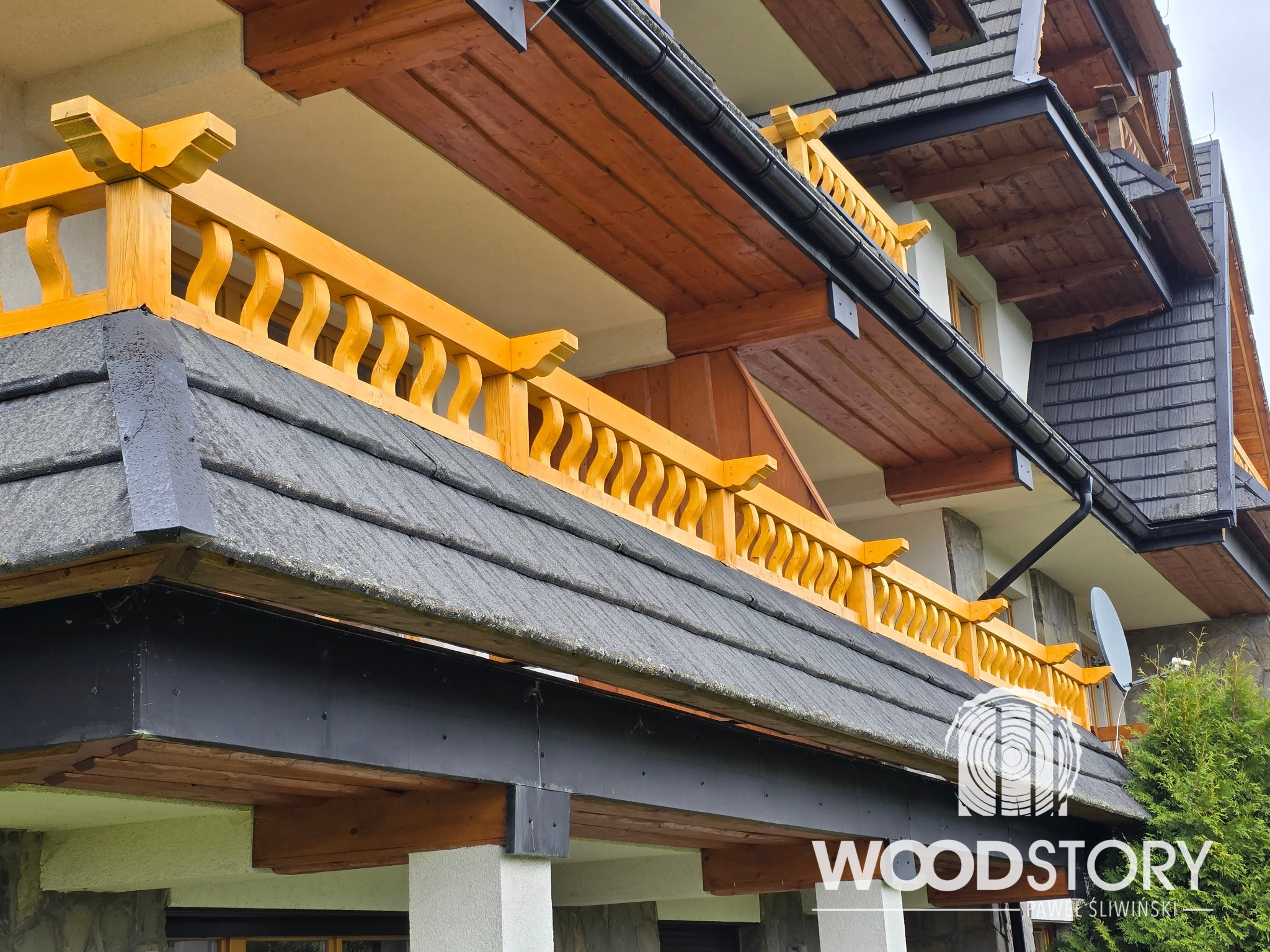 Drewniana balustrada balkonowa w stylu góralskim, pomalowana na żółto, z widocznymi detalami zdobień i fragmentem dachu pokrytego gontem. Ujęcie z dołu.