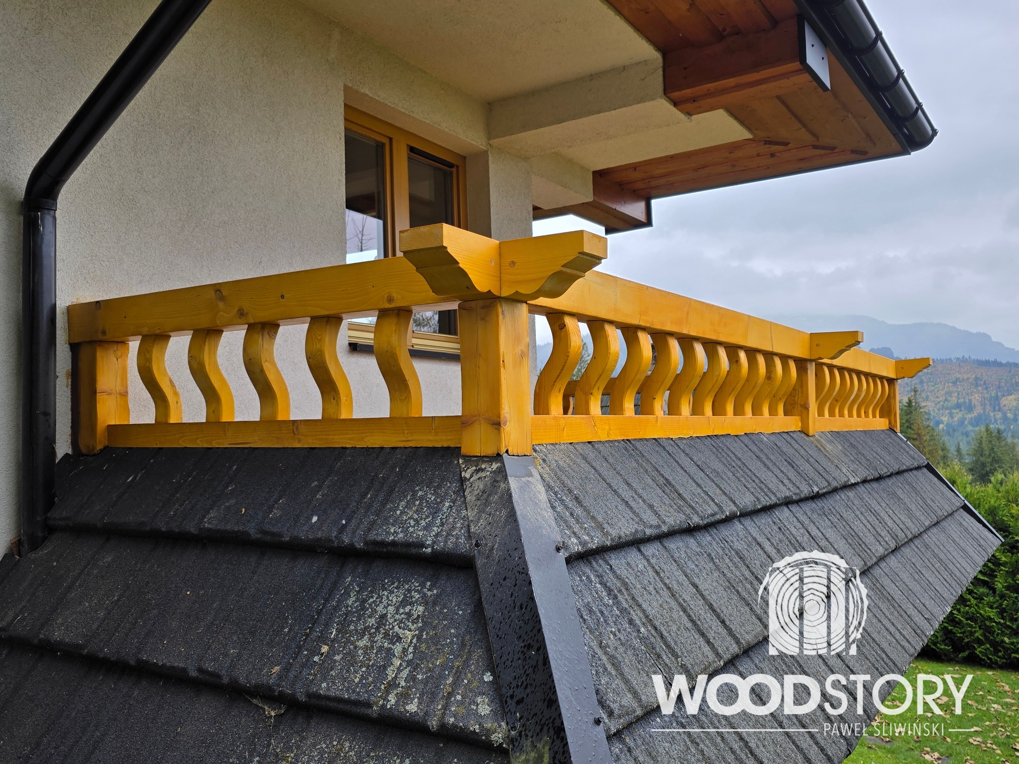 Żółta, drewniana balustrada balkonowa z dekoracyjnymi wycięciami, zamontowana na dachu budynku z czarną dachówką. W tle widok na góry i las.