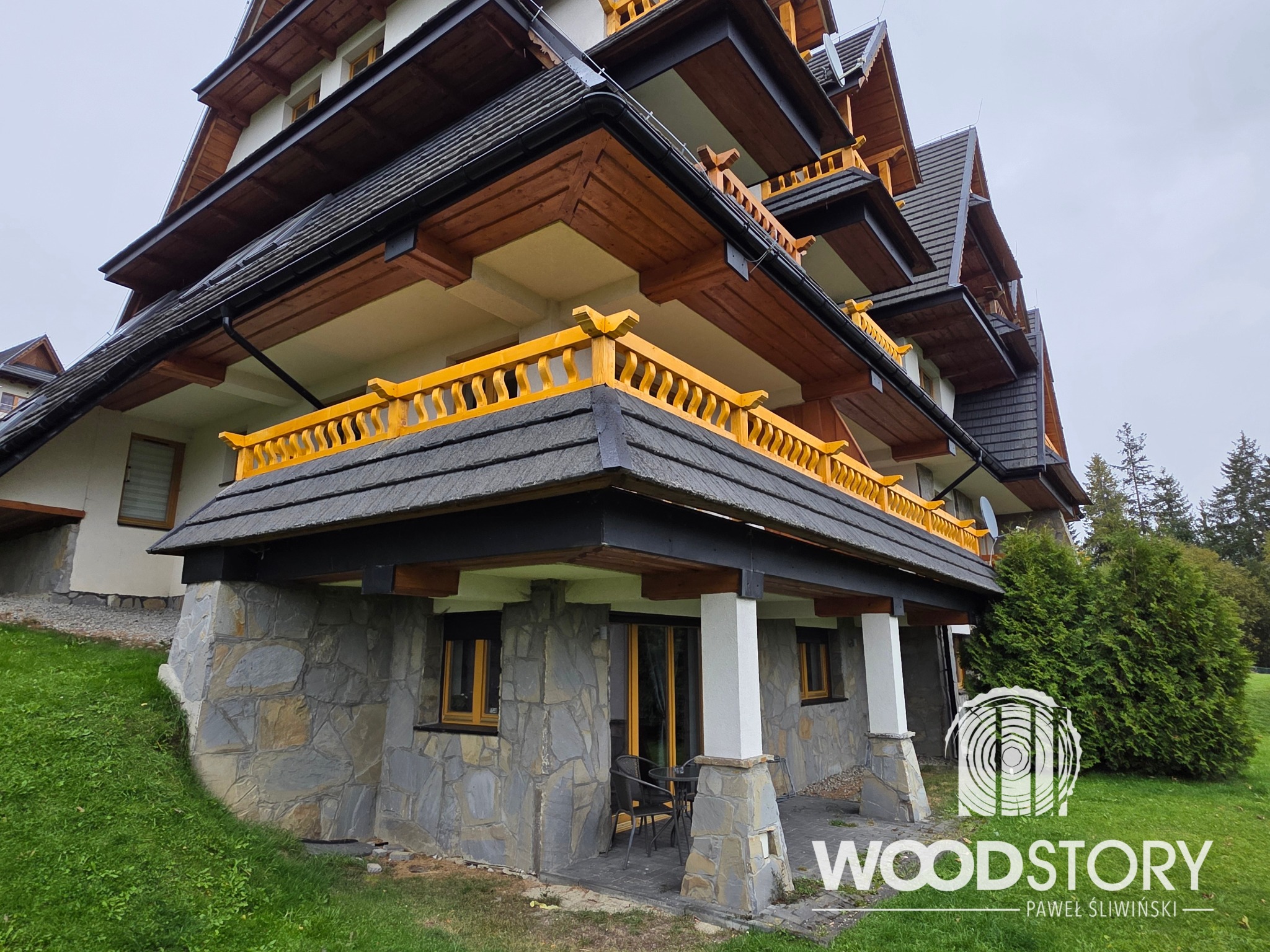 Wielopoziomowy budynek z balkonami o żółtych balustradach, wykończony kamieniem i drewnem, otoczony zielenią. Widoczne detale architektoniczne i naturalne materiały.