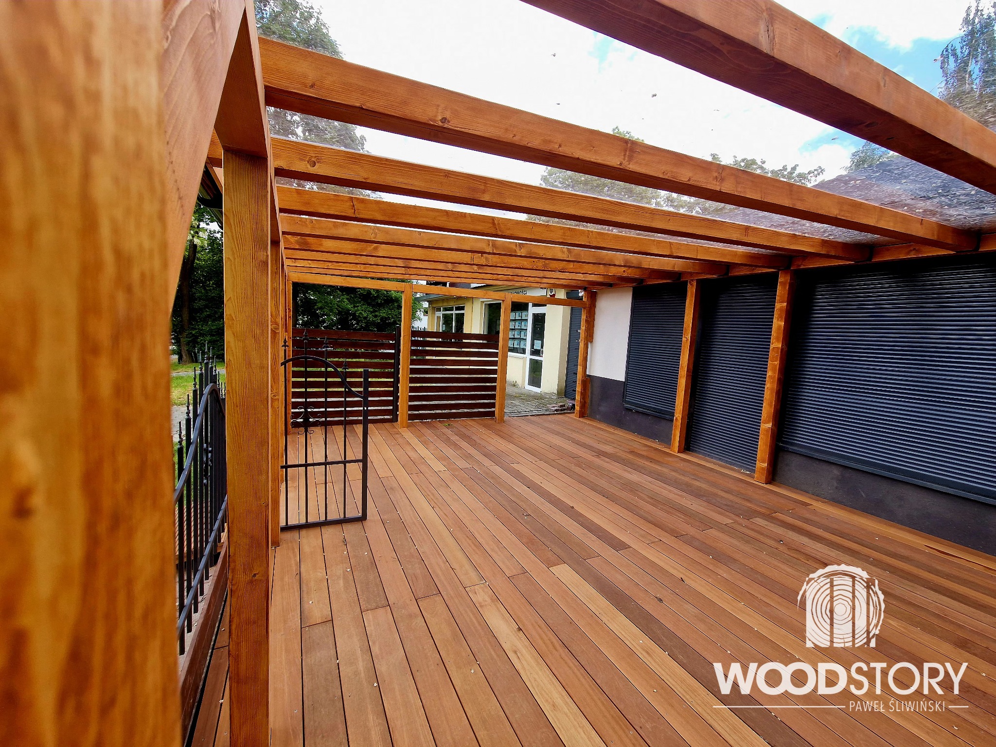 Drewniana pergola z przezroczystym zadaszeniem, widok z dołu, ukazujący konstrukcję i deskowany taras. Metalowa furtka i zieleń w tle. Logo firmy na podłodze.