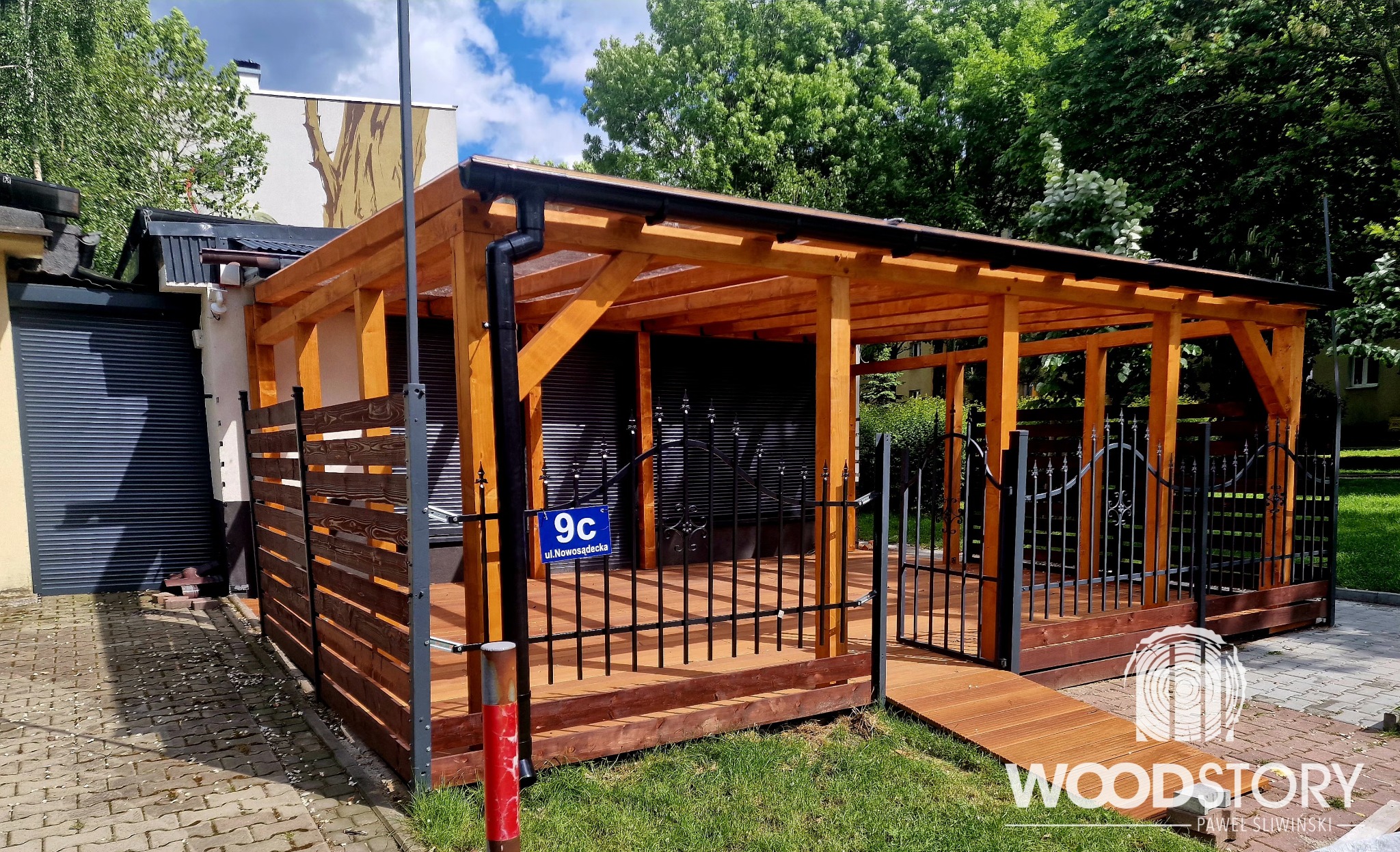 Drewniana pergola z balustradą i bramką z kutego żelaza, zadaszona przezroczystym materiałem, z widocznym adresem na bramce, otoczona zielenią i budynkami.