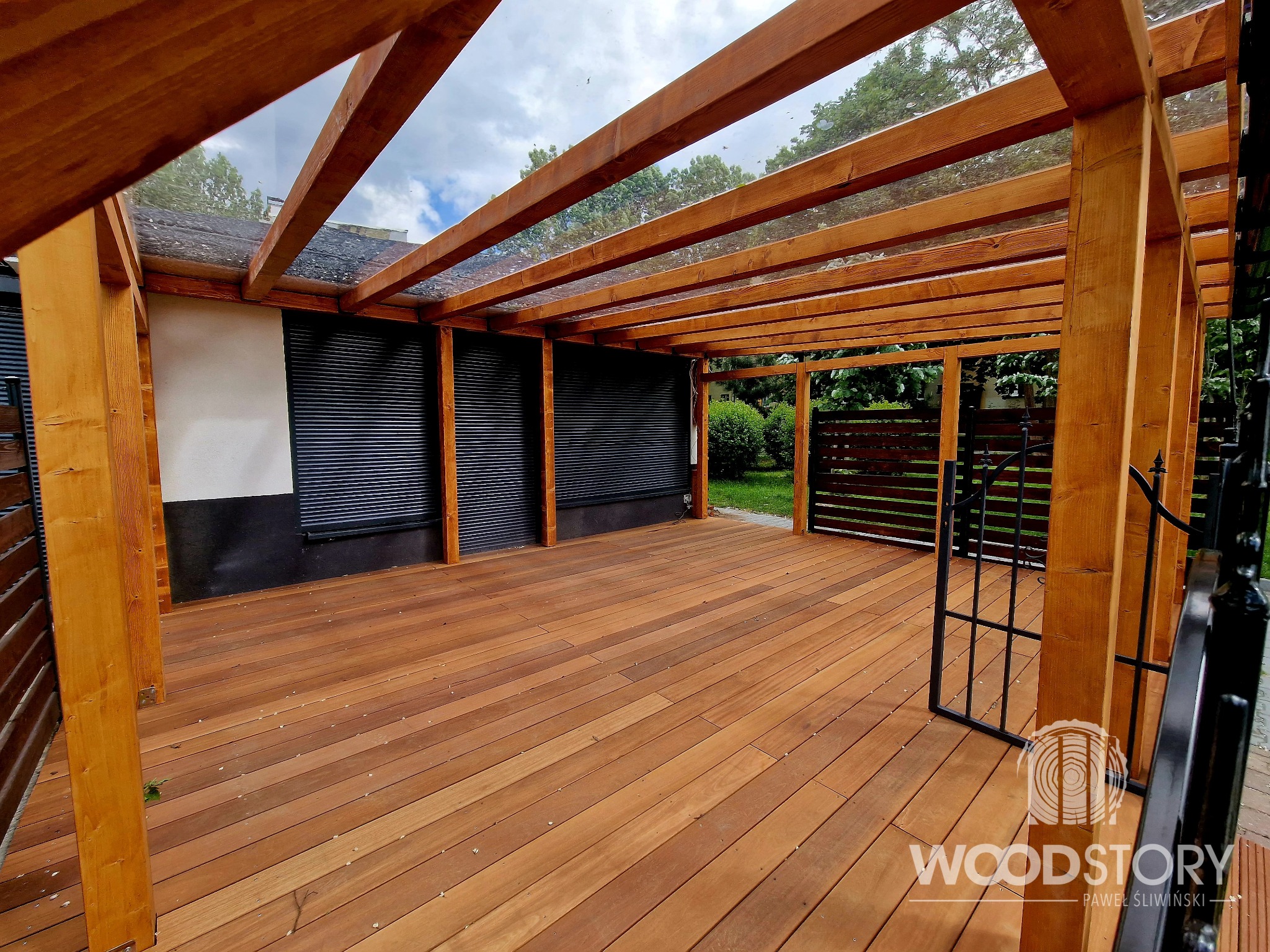 Zadaszona, drewniana konstrukcja tarasowa z przezroczystym dachem, widok na ogród i budynek z żaluzjami. Ciepłe oświetlenie podkreśla naturalny kolor drewna.