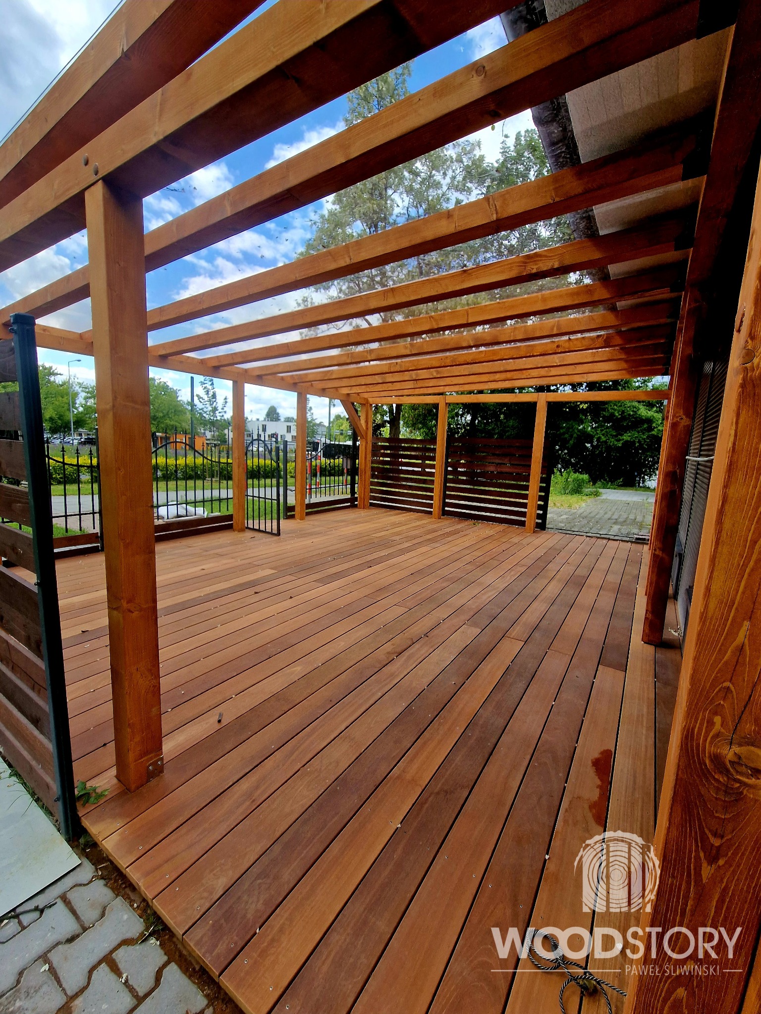 Drewniana pergola z tarasem, widok z dołu, ukazujący konstrukcję z belek i desek. W tle zieleń i błękitne niebo. Na pierwszym planie logo firmy.