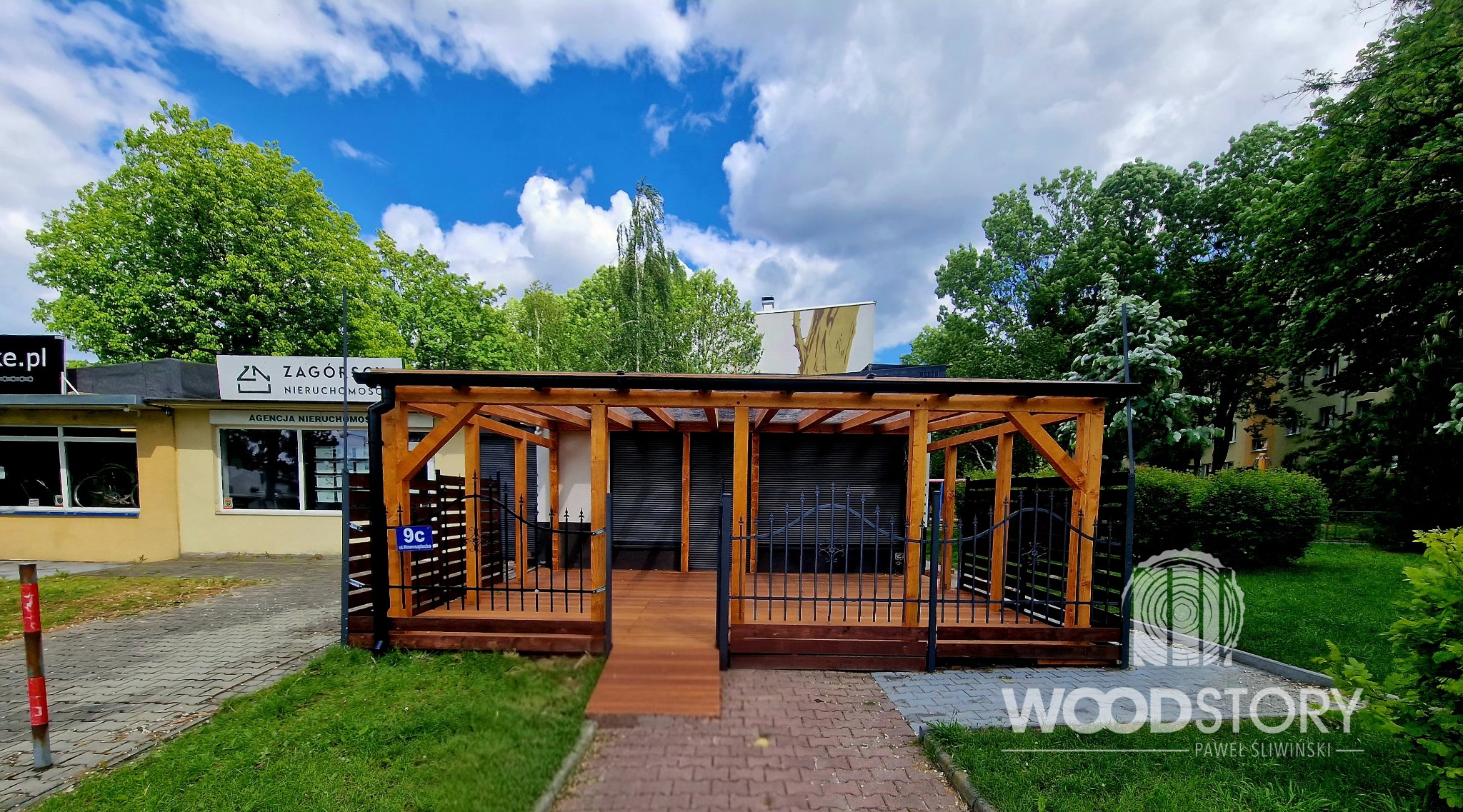 Drewniana pergola z czarnymi, metalowymi elementami, zadaszona, z podłogą z desek, na tle zieleni i błękitnego nieba z chmurami. Widok z zewnątrz.