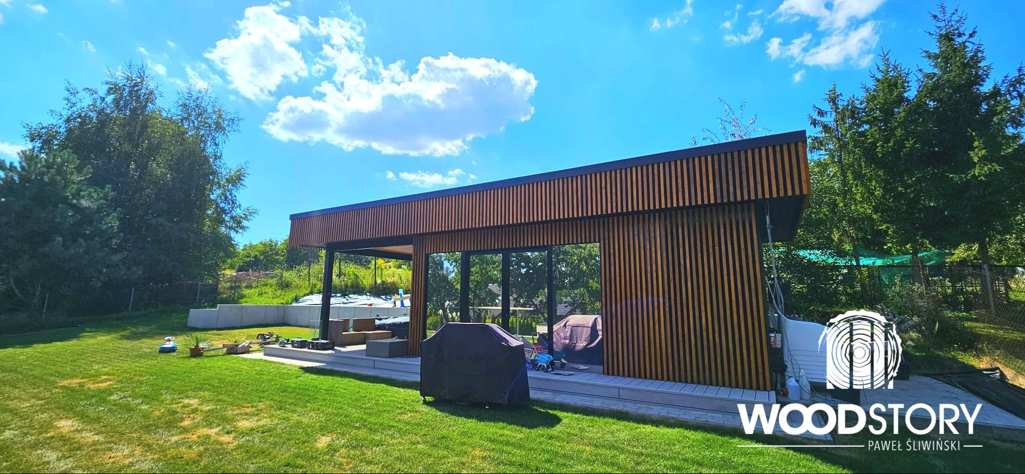 Nowoczesna altana z drewnianymi lamelami i przeszkleniami, zlokalizowana w Wieliczce. Widok na zadbany trawnik i zieleń. Minimalistyczny design i naturalne materiały.