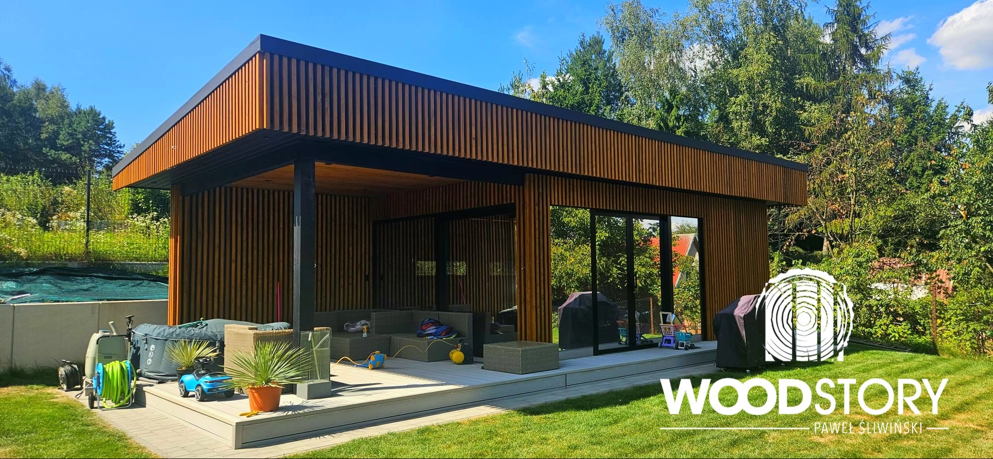 Nowoczesna altana z drewna z przesuwnymi drzwiami, otoczona zielenią. Widoczne meble ogrodowe i akcesoria. Całość utrzymana w ciepłej kolorystyce.