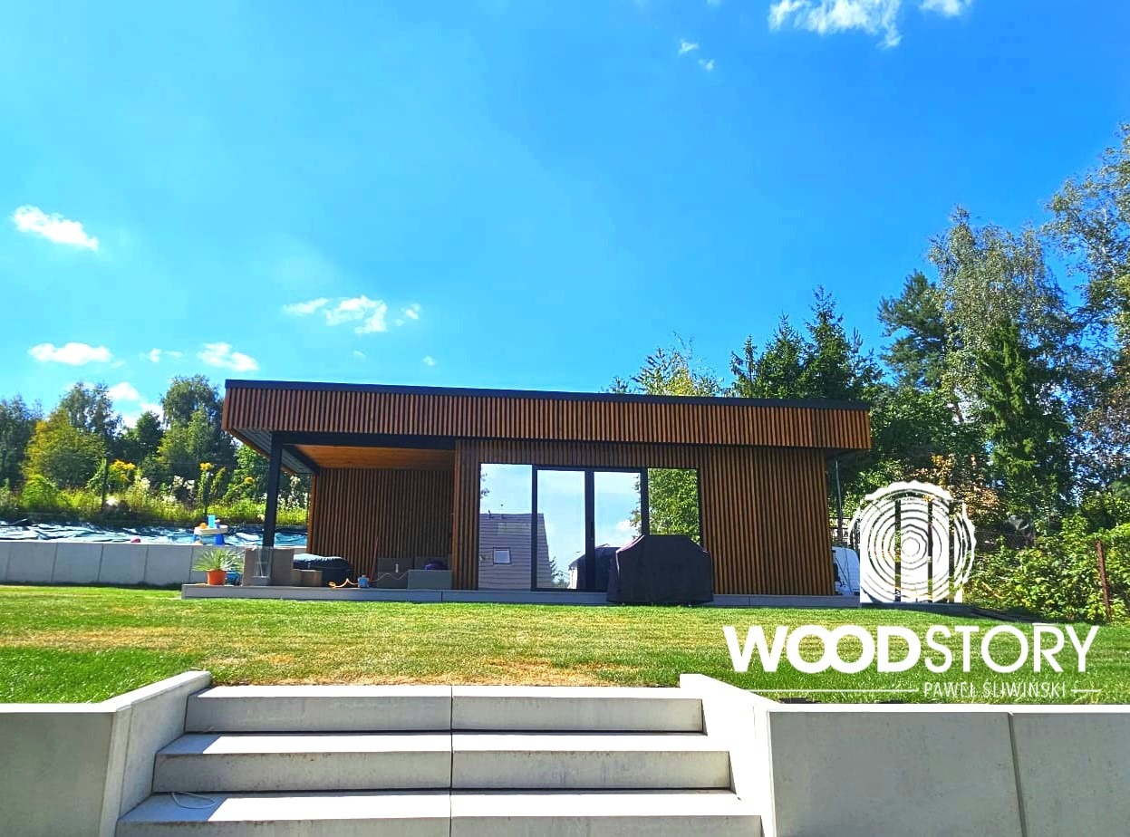 Nowoczesna, drewniana altana z dużymi przeszkleniami, widoczna z perspektywy schodów na trawniku, w tle zieleń drzew i błękitne niebo. Minimalistyczny design.