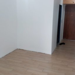 Piotr Maliniak - Pomalowane na biało ściany w rogu pokoju z ułożoną podłogą z płytek imitujących drewno, widoczna lampa sufitowa zabezpieczona folią malarską.