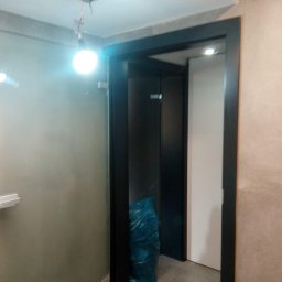 Piotr Maliniak - Wnętrze łazienki w trakcie prac: zamontowane drzwi z czarną ościeżnicą, widoczna kabina prysznicowa z matowego szkła i niebieski worek z odpadami budowlanymi przy wejściu.