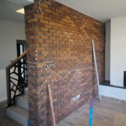 Piotr Maliniak - Ściana z cegły klinkierowej w trakcie montażu, widoczne krzyżyki dystansowe i poziomica, fragment schodów z metalową balustradą w tle.