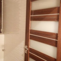 Piotr Maliniak - Zamknięte, drewniane drzwi wewnętrzne z poziomymi, mlecznymi szybami, zamontowane w łazience z beżowymi płytkami o falistej strukturze.