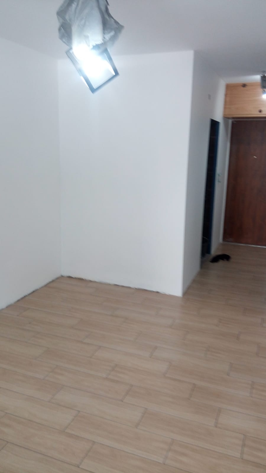 Pomalowane na biało ściany w rogu pokoju z ułożoną podłogą z płytek imitujących drewno, widoczna lampa sufitowa zabezpieczona folią malarską.