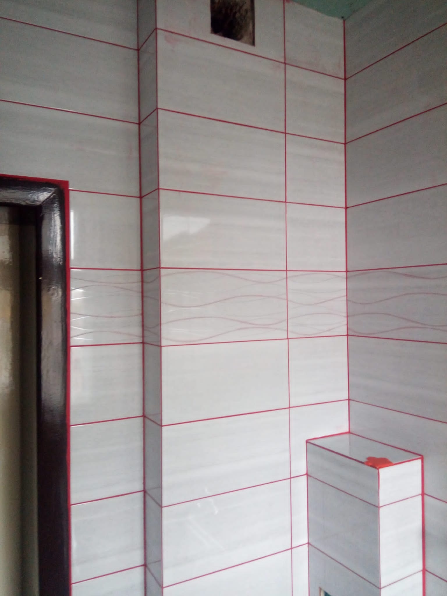 Świeżo położone, białe płytki z czerwonymi fugami w narożniku łazienki, widoczny fragment futryny drzwi.