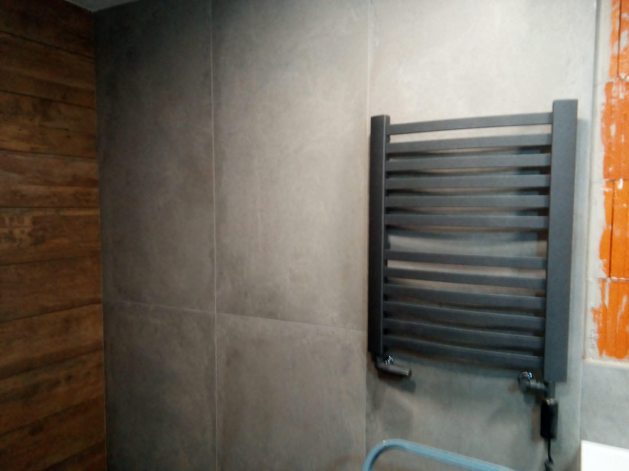 Szary grzejnik drabinkowy zamontowany na ścianie z szarych płytek w łazience, widoczny fragment ściany z drewnianą okładziną oraz ściana w trakcie wykończenia z widocznym klejem do płytek.