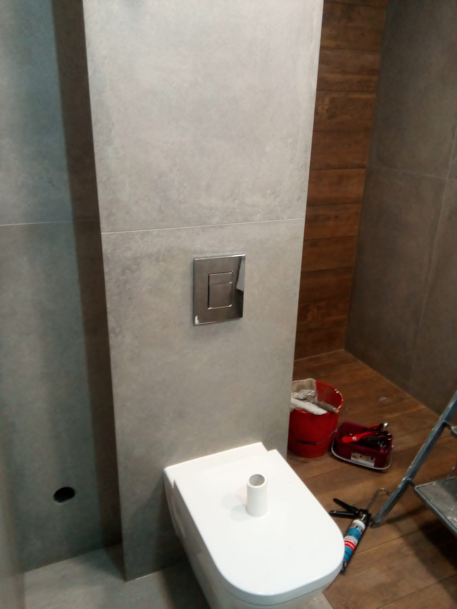 Świeżo zamontowana, podwieszana toaleta z chromowanym przyciskiem spłukującym, w łazience z szarymi i drewnopodobnymi płytkami, widoczny kubeł z narzędziami i drabina.