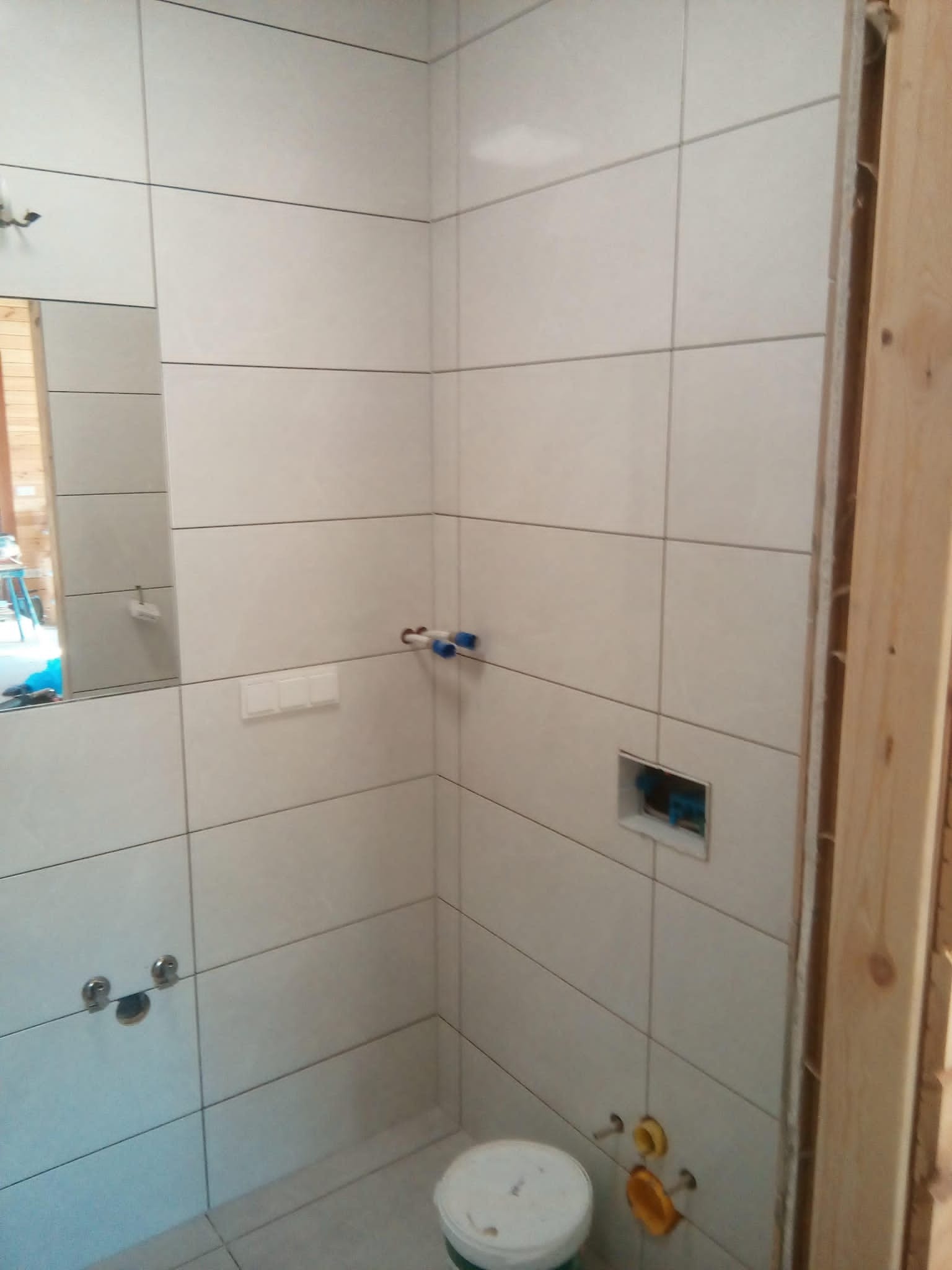 Wnętrze łazienki w trakcie remontu, widoczne białe płytki na ścianach, przygotowane instalacje wodne i elektryczne, oraz fragment nieobudowanej ramy drzwiowej.