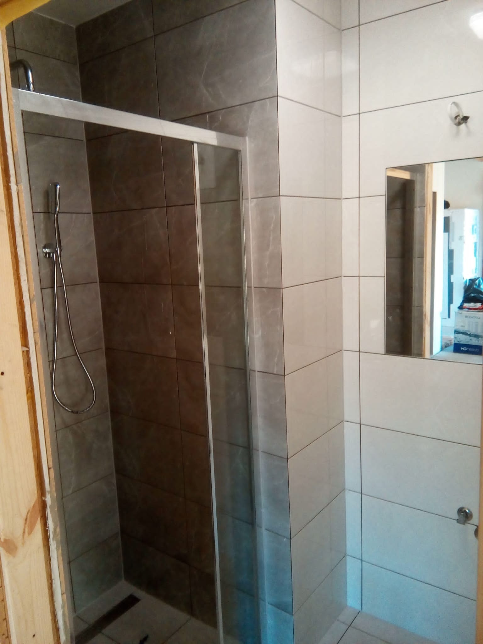 Nowoczesna kabina prysznicowa z przesuwnymi drzwiami i szarymi płytkami w trakcie montażu, widoczne surowe drewno ościeżnicy.