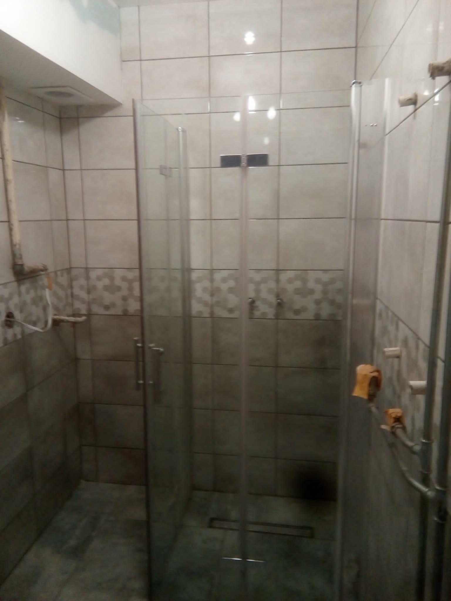 Szklana kabina prysznicowa z dwuskrzydłowymi drzwiami zamontowana w łazience wyłożonej szarymi płytkami w dwóch odcieniach, widoczne rury instalacji wodnej.