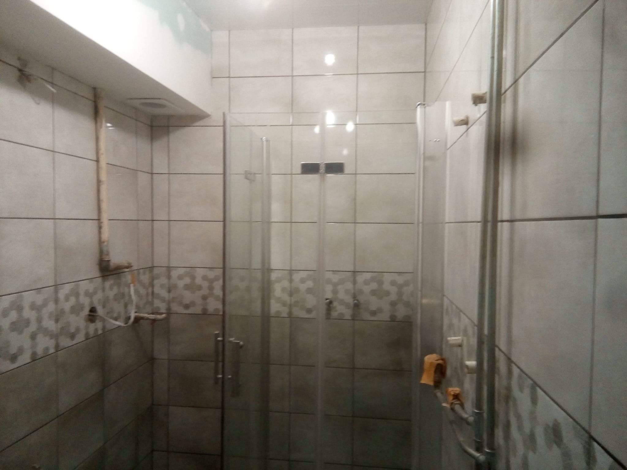 Nowoczesna kabina prysznicowa ze szklanymi drzwiami, wykończona szarymi płytkami z dekoracyjnym pasem wzorzystych płytek na ścianach, widoczne rury instalacyjne i żółta ściereczka.