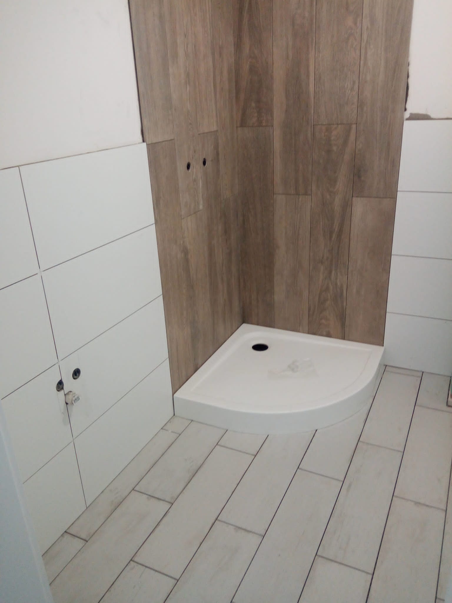 Narożny brodzik z odpływem, obłożony płytkami imitującymi drewno na ścianie i podłodze w trakcie prac wykończeniowych łazienki, widoczne niedokończone połączenia płytek.