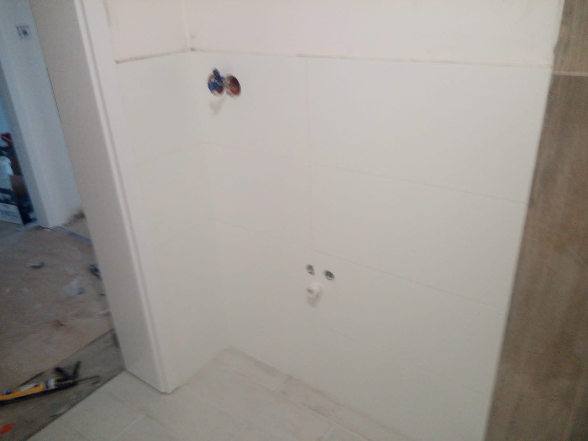 Ściana łazienki w trakcie wykończenia: białe płytki ułożone poziomo, widoczne przyłącza wodne i otwory na baterię, podłoga częściowo wyłożona płytkami, w tle widoczny bałagan remontowy.