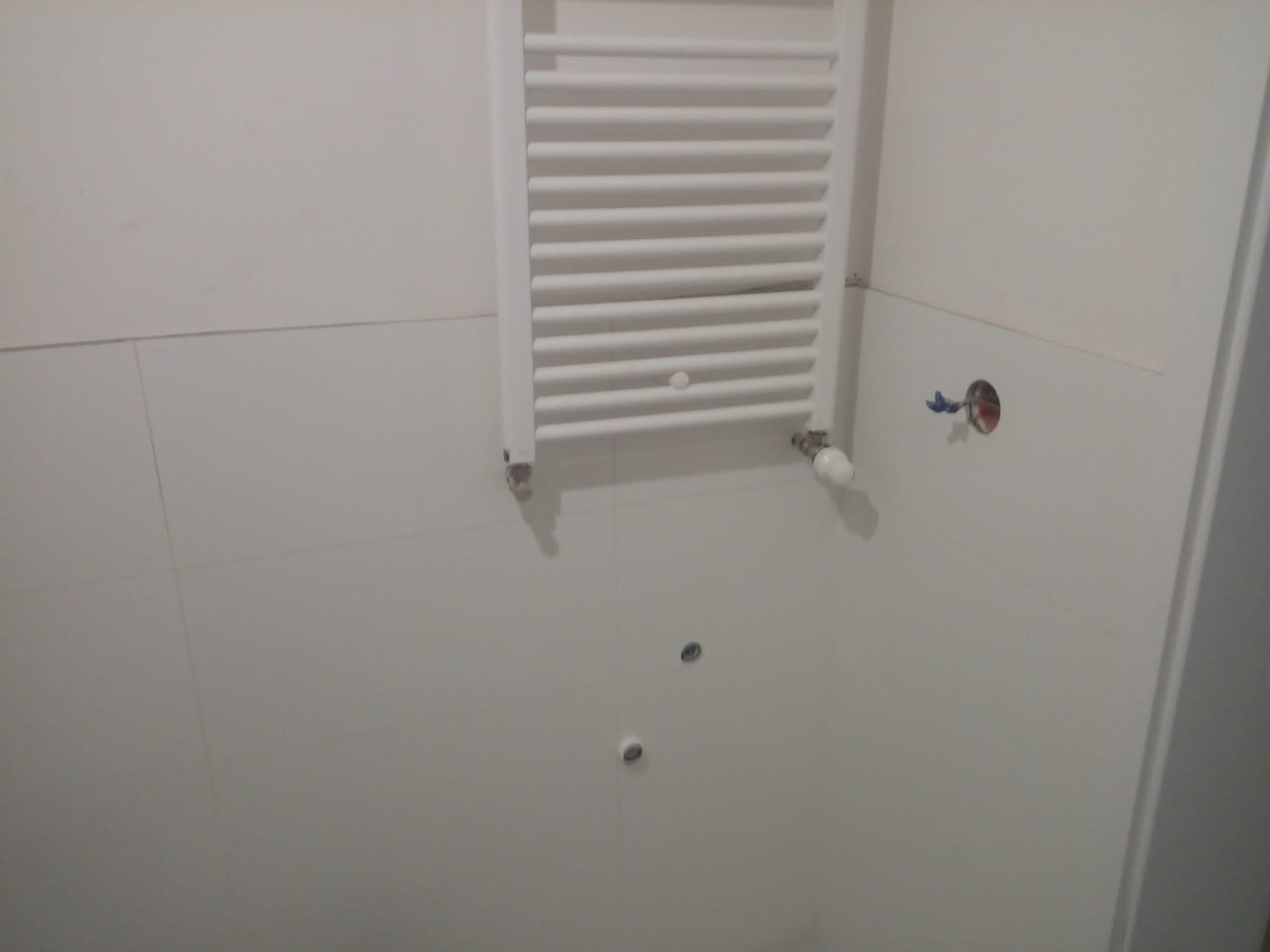 Biały, drabinkowy grzejnik łazienkowy zamontowany na ścianie z jasnych płytek, widoczne przygotowane otwory pod instalację.