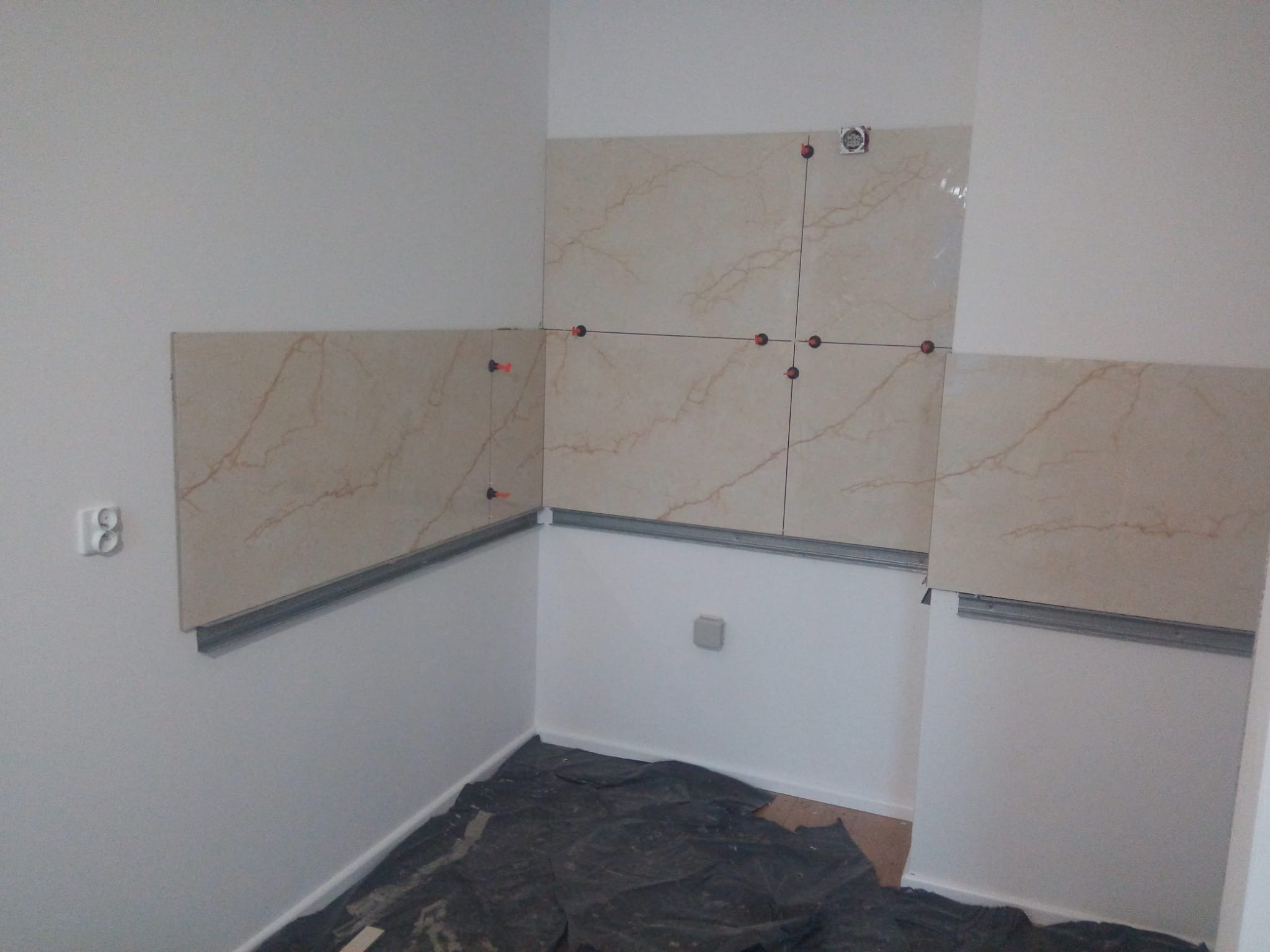 Układanie beżowych płytek imitujących marmur na ścianie w rogu pomieszczenia, widoczne listwy i krzyżyki dystansowe, podłoga zabezpieczona folią.