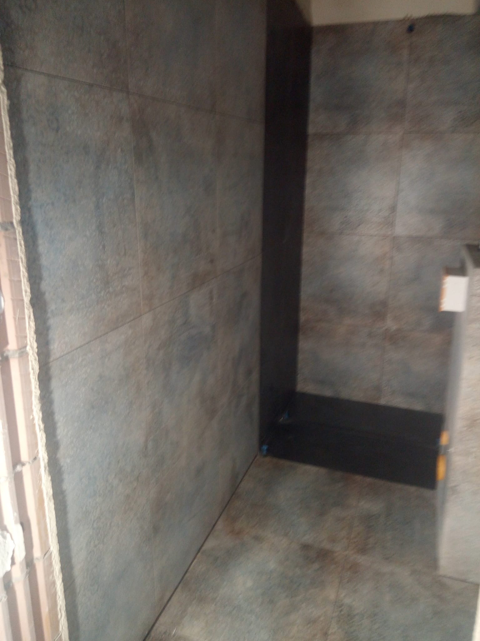 Wnętrze łazienki w trakcie prac wykończeniowych, widoczne szare płytki na ścianach i podłodze, fragment ściany z cegieł oraz czarny element brodzika.