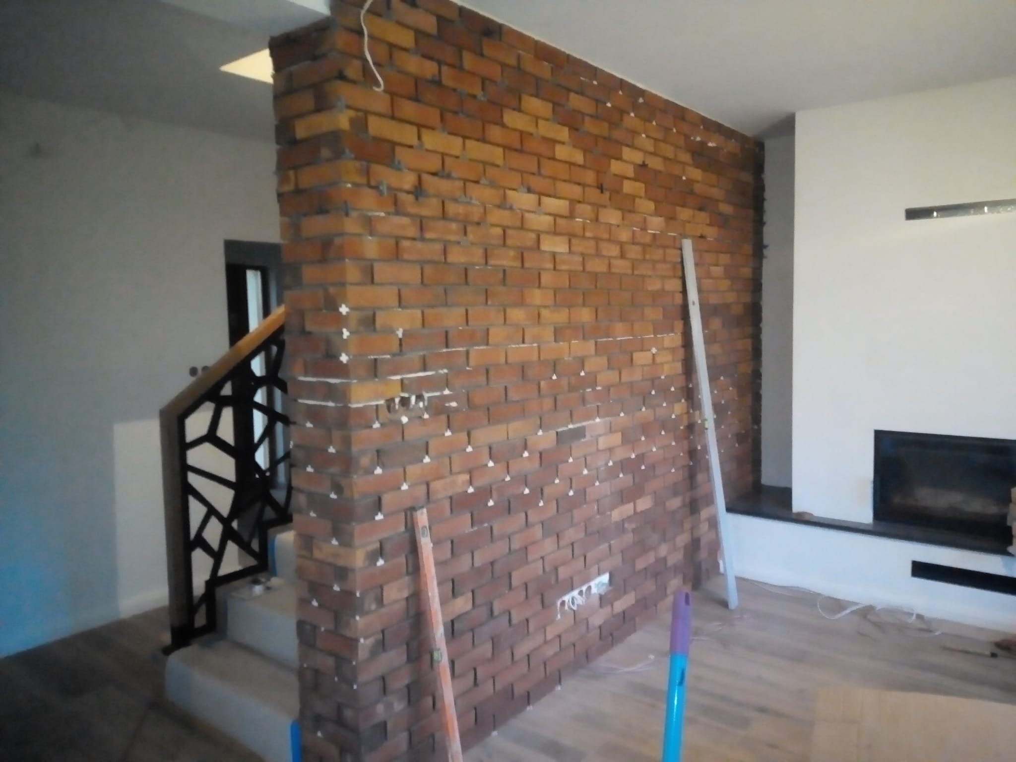 Ściana z cegły klinkierowej w trakcie montażu, widoczne krzyżyki dystansowe i poziomica, fragment schodów z metalową balustradą w tle.