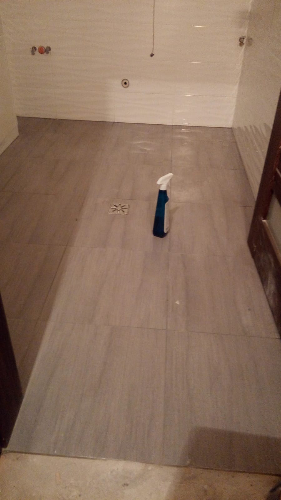Wykończona podłoga w łazience z szarych, matowych płytek, widoczny odpływ liniowy i butelka z rozpylaczem do czyszczenia.