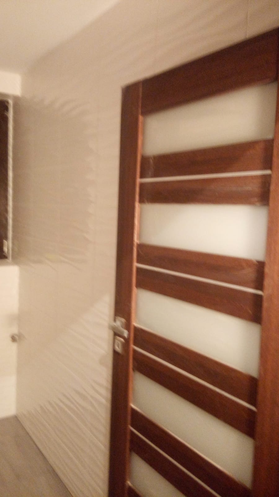 Zamknięte, drewniane drzwi wewnętrzne z poziomymi, mlecznymi szybami, zamontowane w łazience z beżowymi płytkami o falistej strukturze.