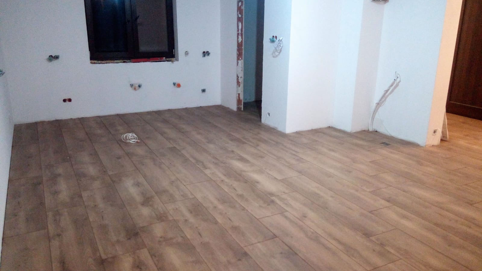 Ułożona podłoga z paneli imitujących drewno w trakcie prac wykończeniowych, widoczne wystające kable i niezabezpieczone gniazdka elektryczne na ścianach.