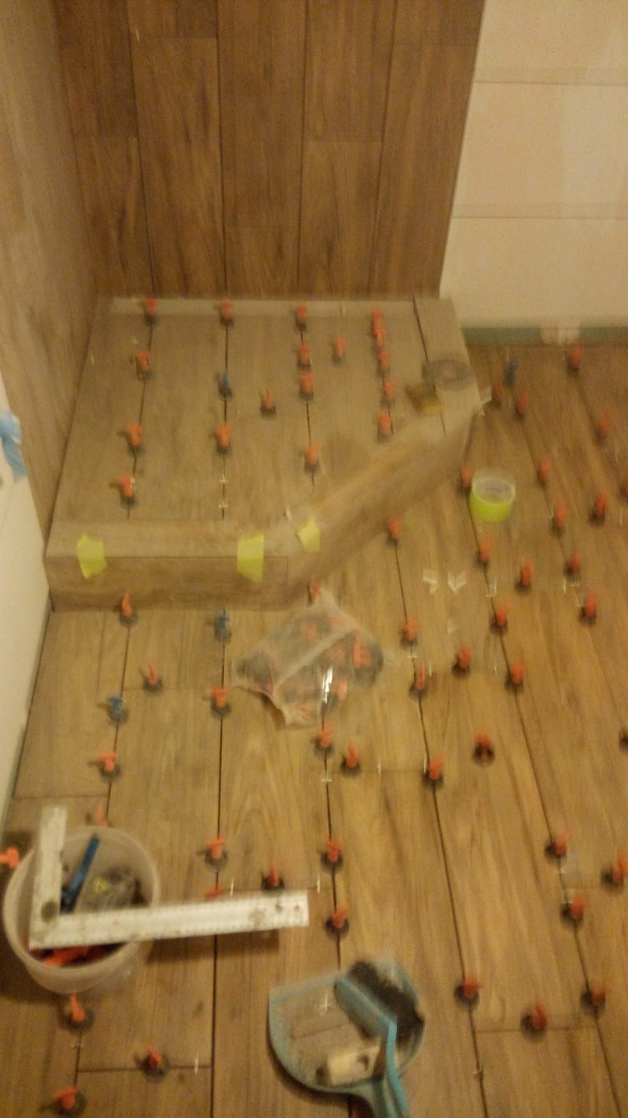 Układanie płytek imitujących drewno na podłodze i schodku w łazience, z użyciem krzyżyków dystansowych i poziomicy, widoczny także pojemnik z narzędziami i paczka z krzyżykami.