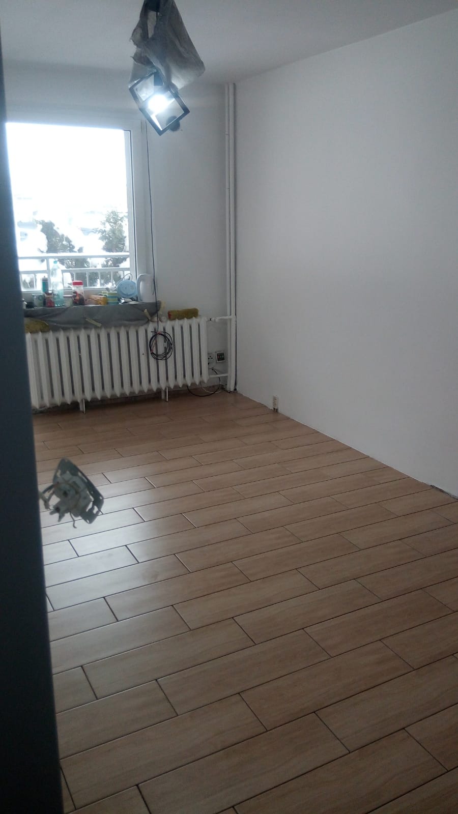 Pokój po remoncie z nową podłogą z płytek imitujących drewno, widoczny grzejnik pod oknem i lampa budowlana zawieszona pod sufitem.