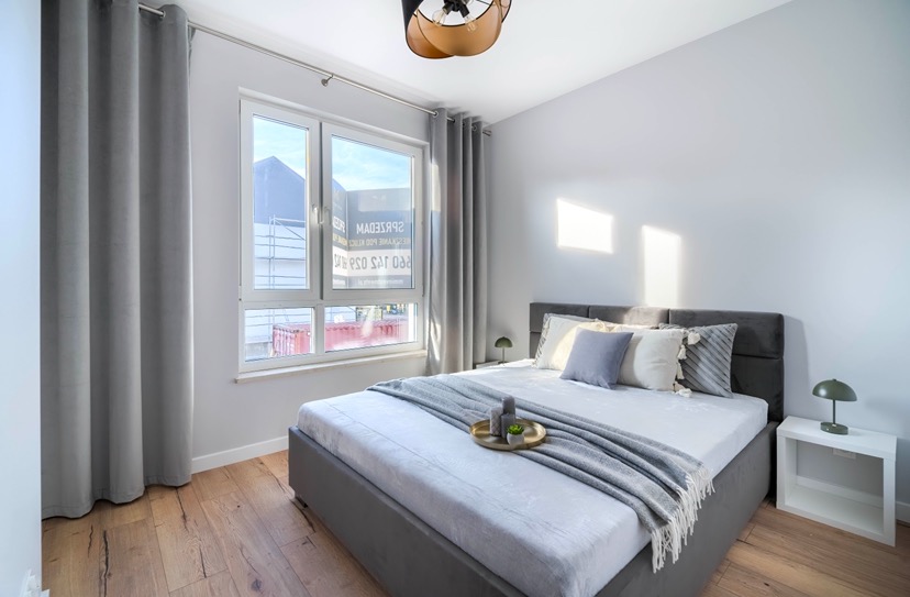 Jasne wnętrze sypialni z szarym łóżkiem, drewnianą podłogą i oknem z widokiem na rusztowania budowlane za oknem.