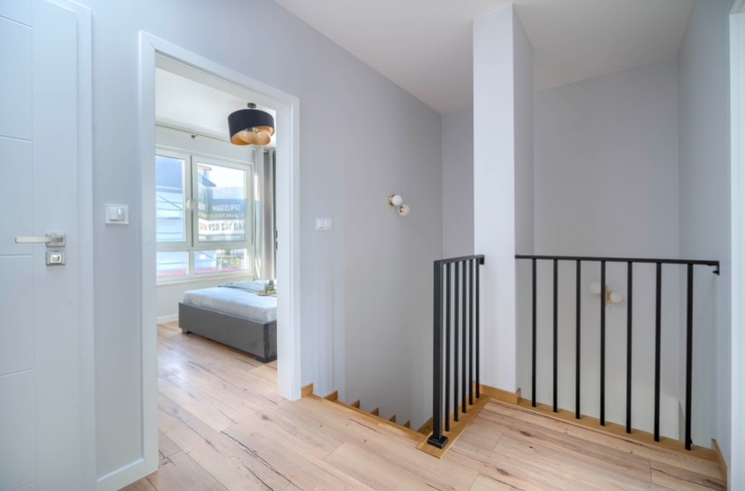Korytarz na piętrze z drewnianą podłogą, białe ściany pomalowane na szaro, czarna balustrada schodów i widok na pokój przez otwarte drzwi.