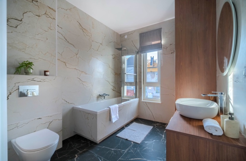 Elegancka łazienka z płytkami imitującymi marmur na ścianach i podłodze, biała wanna z hydromasażem, umywalka nablatowa na drewnianej szafce i okrągłe lustro.