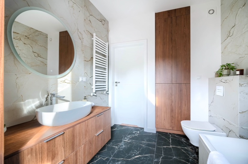 Nowoczesna łazienka z owalnym lustrem podświetlanym od tyłu, marmurowymi płytkami na ścianach, szafką z drewnopodobnym fornirem pod umywalką nablatową i czarną marmurową posadzką.