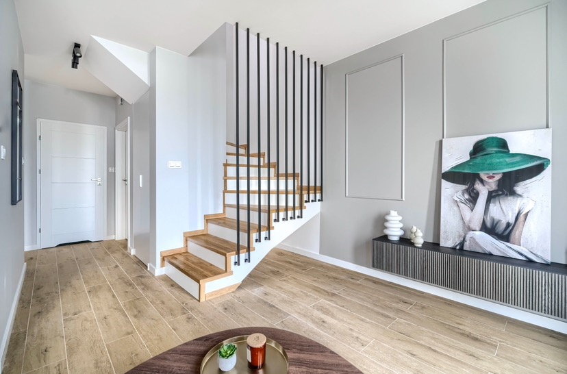Jasne wnętrze z drewnianymi schodami z czarnymi poręczami, obrazem kobiety w zielonym kapeluszu i podłogą imitującą drewno.