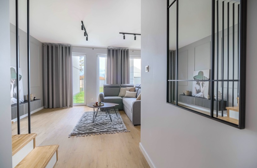 Jasne wnętrze salonu z drewnianą podłogą, szarą sofą narożną, dywanem w geometryczne wzory i czarnymi reflektorami sufitowymi. W tle widoczne okna z szarymi zasłonami i obraz w czarnej ramie.