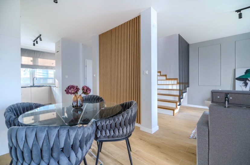 Nowoczesne wnętrze z okrągłym szklanym stołem otoczonym szarymi, tapicerowanymi krzesłami, drewnianą ścianką działową z pionowych listew, jasną kuchnią w tle oraz schodami z czarną balustradą.